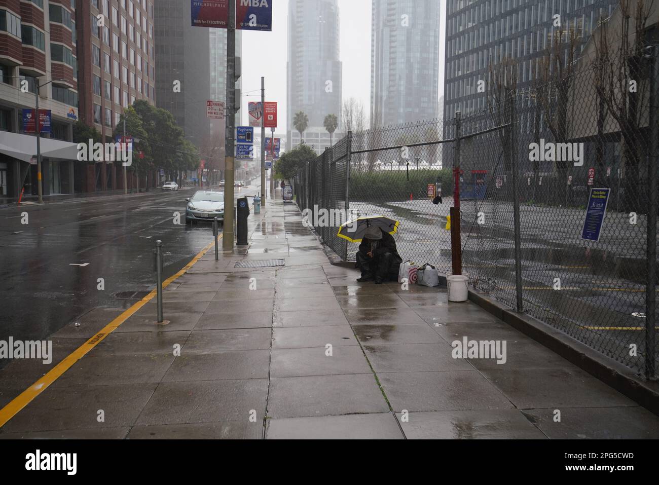 A person sheltering under an umbrella on a rain-soaked San Francisco sidewalk beside a construction fence